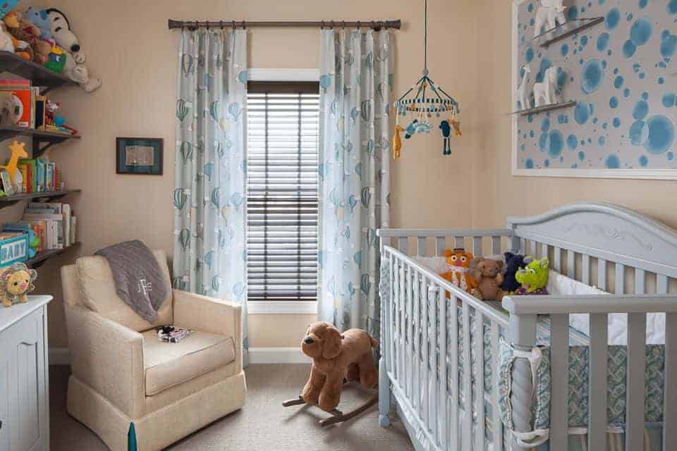 A cute baby mobile hangs over the gray crib in this beige nursery offering a skirted armchair and wooden floating shelves filled with books and toys.