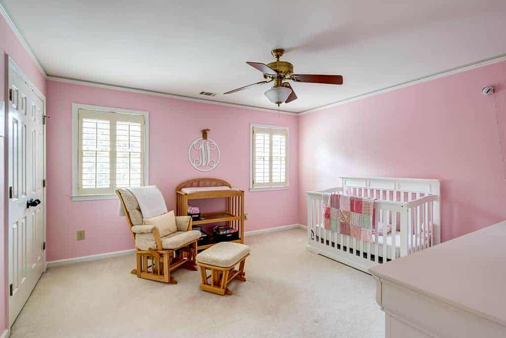 Pink nursery with a white crib and wooden shelving complementing with the cushioned armchair and matching ottoman. It has a classic ceiling fan and white framed windows enclosed in cream shutters.