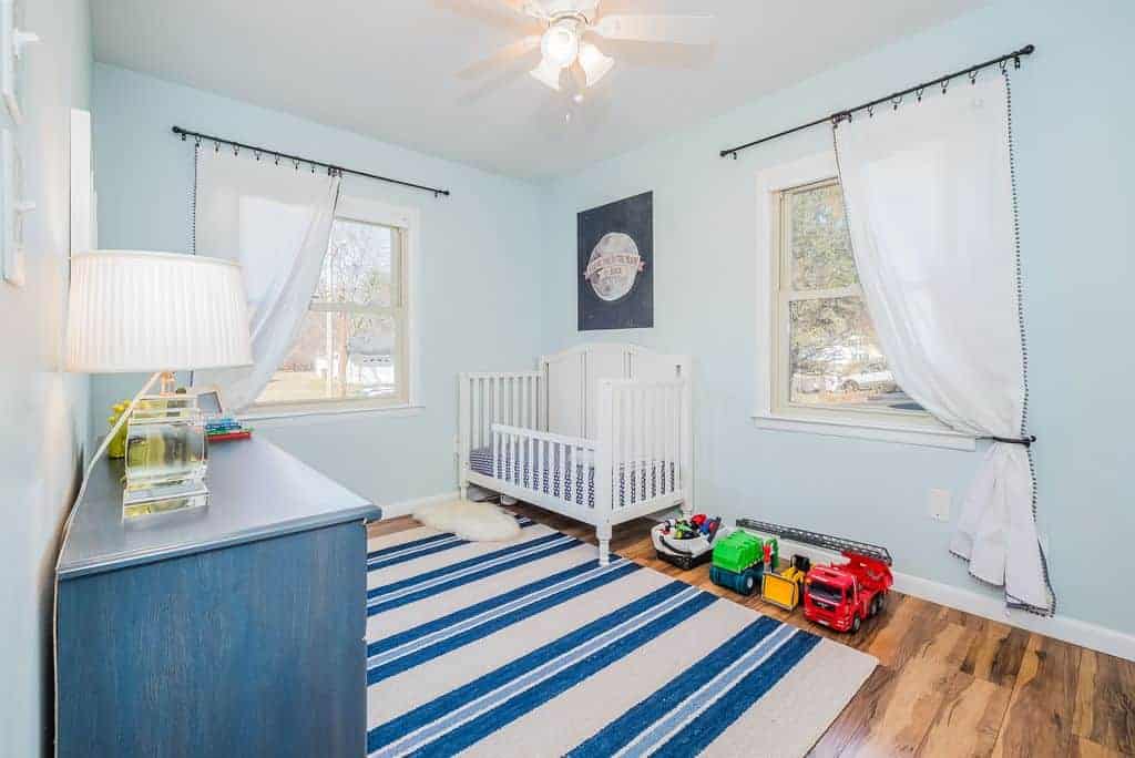 Bright nursery boasts a white crib and blue cabinet topped by a table lamp with pleated shade. It includes a white ceiling fan with lights and a striped rug that lays on the hardwood flooring.