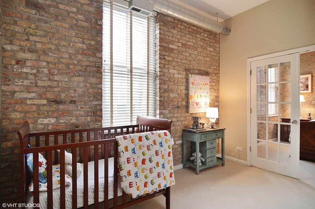 A stone brick accent wall adds texture in this industrial nursery with carpet flooring and a French door that leads out to a bedroom. It includes a wooden crib and distressed cabinet with a table lamp and artwork on top.