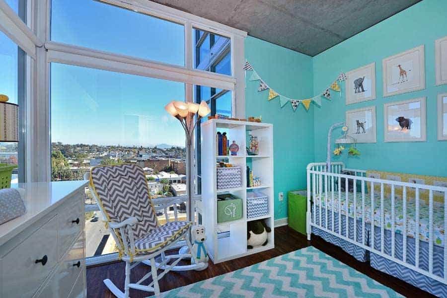 This nursery is decorated with garland flag and animal artworks mounted above the skirted crib. It has a chevron rug and armchair against the full height glazing overlooking the incredible city view.