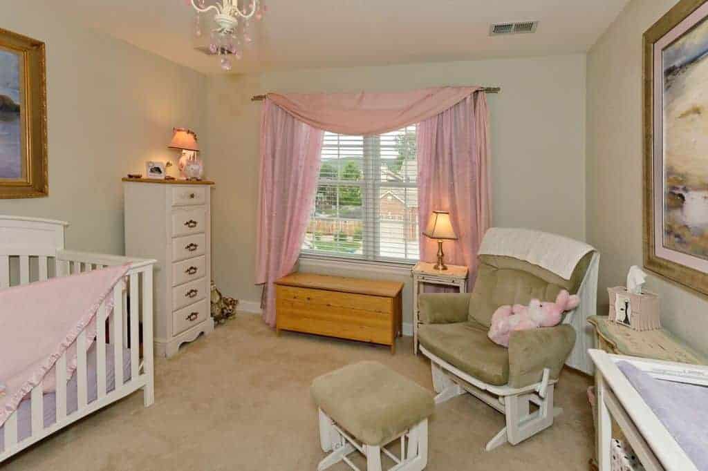 Beige nursery with subtle pink accents from the drapes, blanket and bunny that sits on a cushioned armchair matching with the ottoman and carpet flooring.