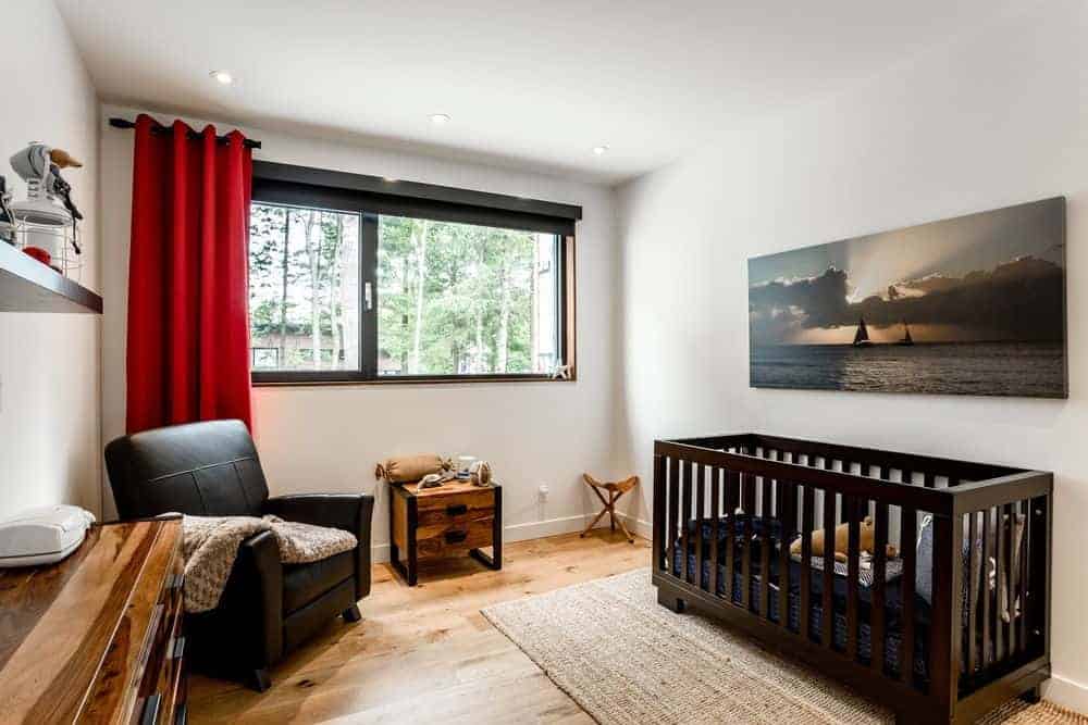 Cozy nursery with a black leather armchair and an ocean canvas mounted above the crib that sits on a jute rug. It has natural hardwood flooring and aluminum framed windows covered in red drapery.