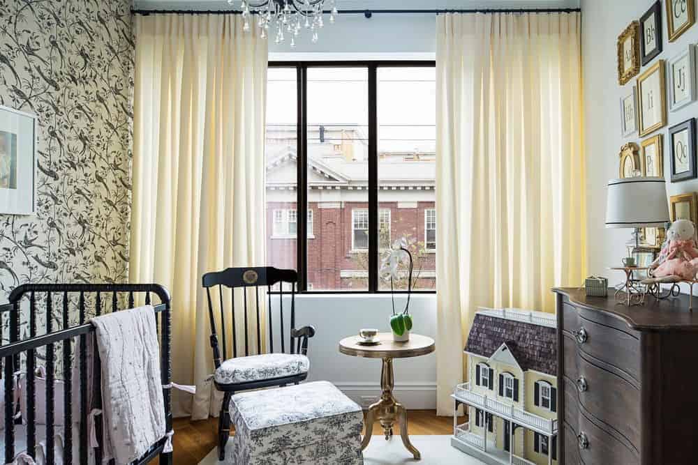 Classy nursery designed with a bird wallpaper and an alphabet letter gallery mounted above the wooden drawer chest. It has a black crib and cushioned chair accompanied by a skirted ottoman and round side table.