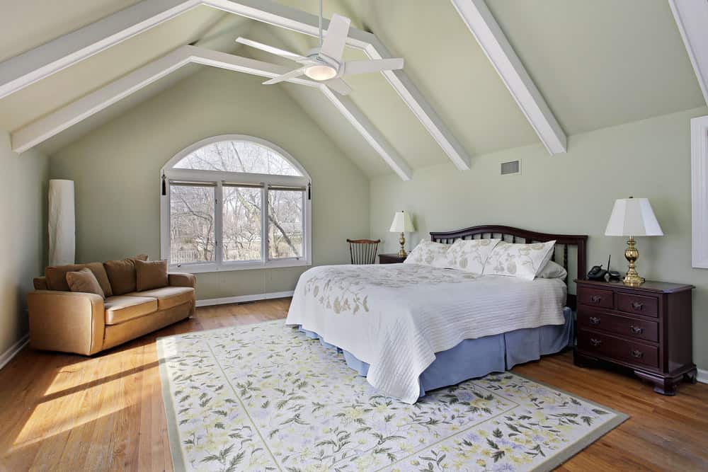 Spacious primary bedroom featuring hardwood flooring and a vaulted ceiling with beams. It offers a large bed along with a brown couch in the corner.