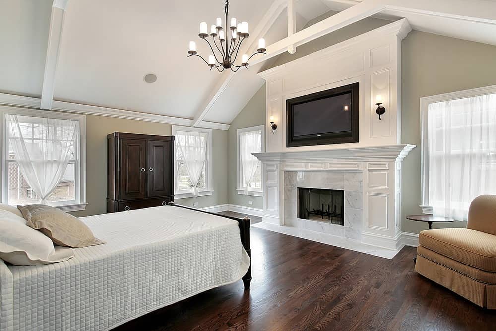 Primary bedroom featuring a tall vaulted ceiling along with hardwood flooring. There's a fireplace and a large TV on the wall.