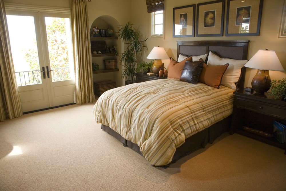 Spacious primary bedroom featuring a classy bed setup along with built-in shelving on the side. The room features a tall ceiling and carpeted flooring.