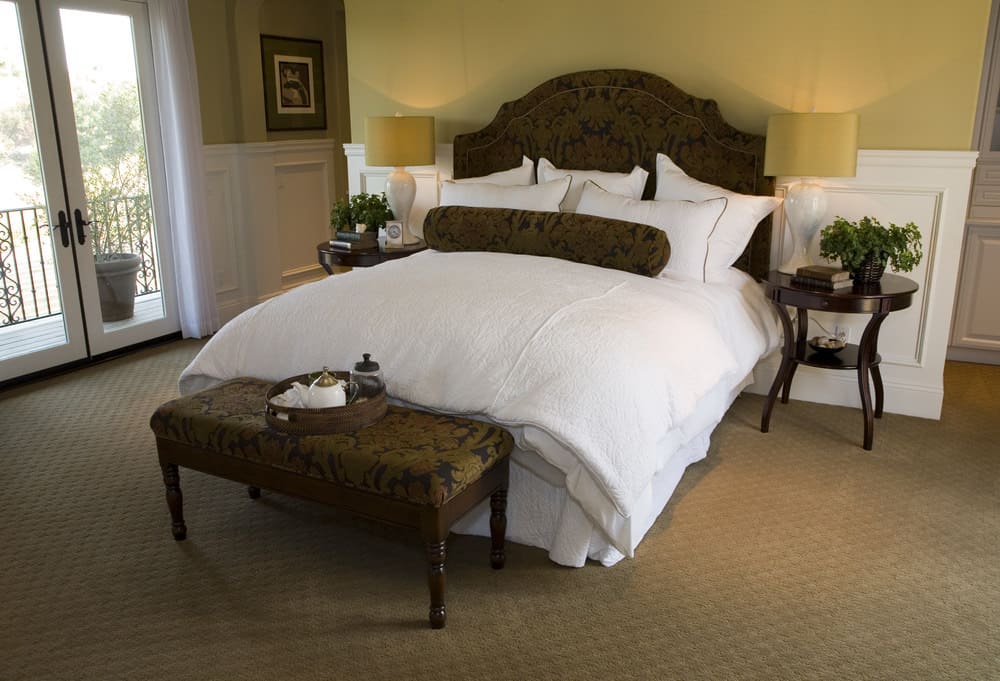 Large primary bedroom featuring a large classy bed set on the room's carpet flooring and is surrounded by yellow walls.
