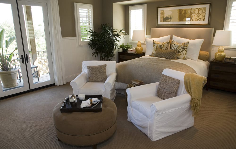 Large primary bedroom featuring carpet flooring and brown walls. The room features a nice classy bed along with a sitting area featuring a brown ottoman.