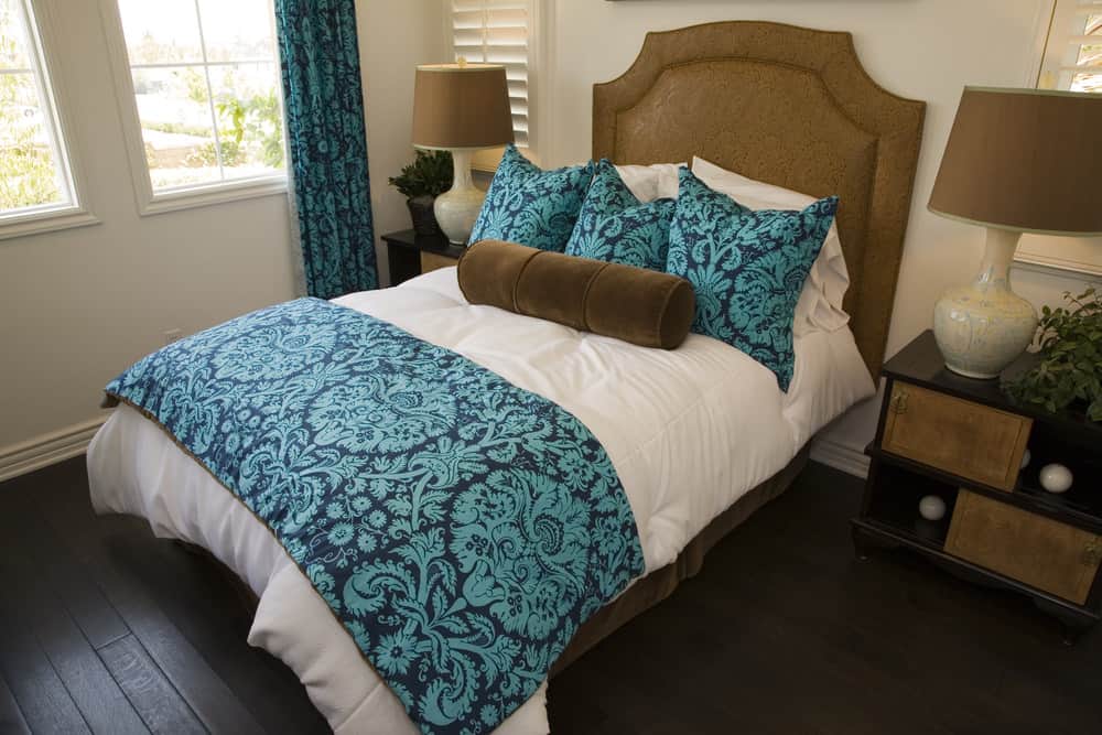 A focused shot at this primary bedroom's classy double-sized bed with a blue accent lighted by brown table lamps matching the brown bed frame.