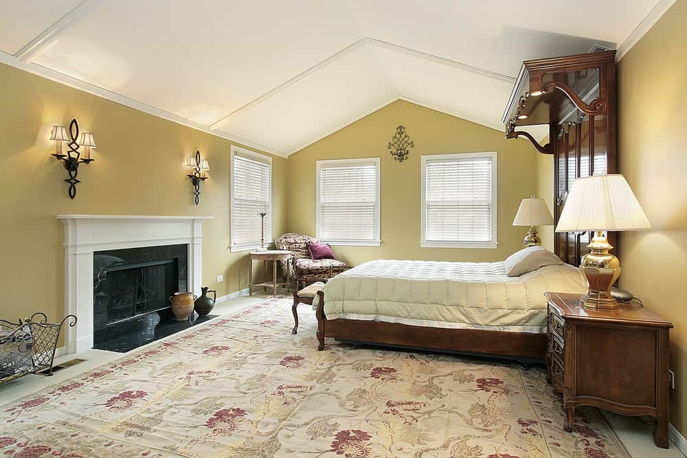Large primary bedroom featuring a vaulted ceiling and a large classy area rug. It has a luxurious large bed along with a large fireplace lighted by wall lights.