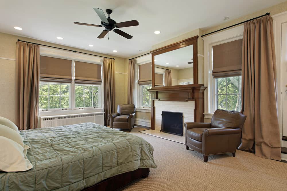 Primary bedroom featuring brown carpet flooring along with brown window curtains. The room offers two brown leather seats along with a fireplace.
