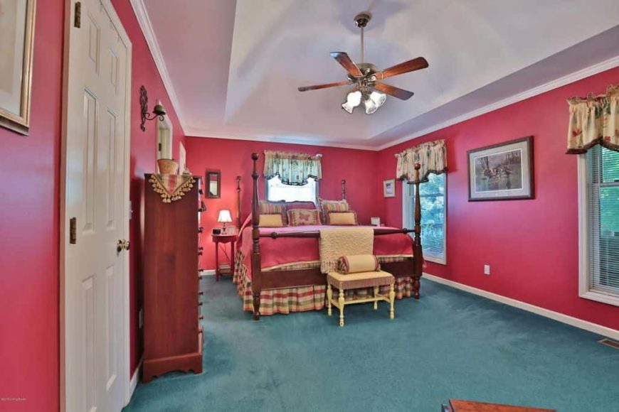 Country style primary bedroom featuring red walls and green carpet flooring. It offers a large classy bed along with cabinetry on the side.