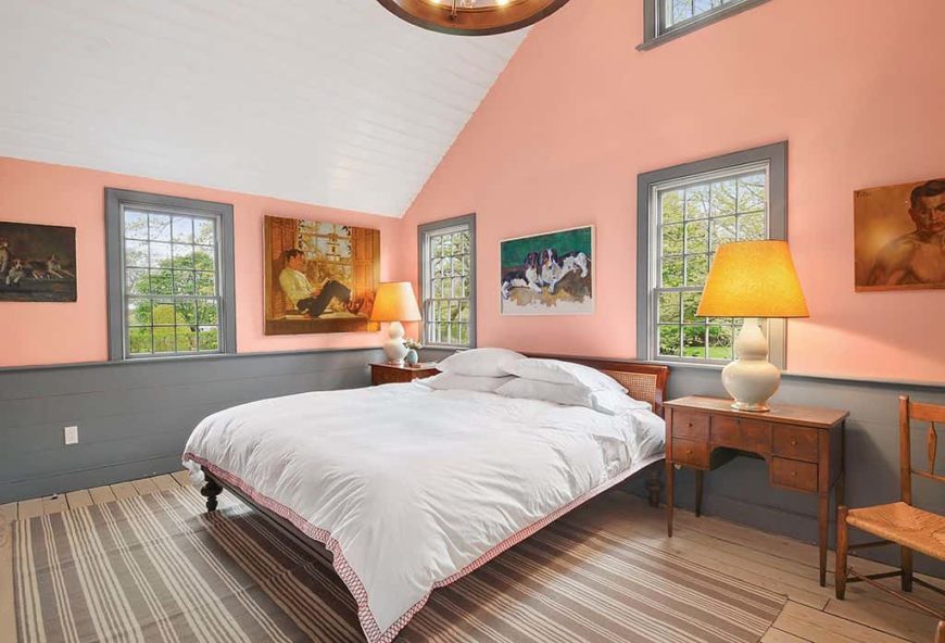 Country style primary bedroom featuring orange walls and hardwood flooring topped by an area rug, along with a tall white ceiling. The bed is lighted by table lamps on both sides.