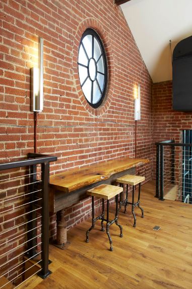 The red brick wall of this industrial-style foyer has a built-in table with water pipes as legs. This is paired with a couple of wooden stools with pipes for legs as well. This is given a nice background of a circular window flanked by modern wall lamps.