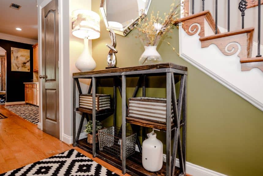 This charming foyer has an industrial-style console table with gray metal rods that pairs well with the wooden shelves filled with decors and supports a table lamp above that casts yellow light on the green wall that complements the hardwood flooring and the steps of the stairs.