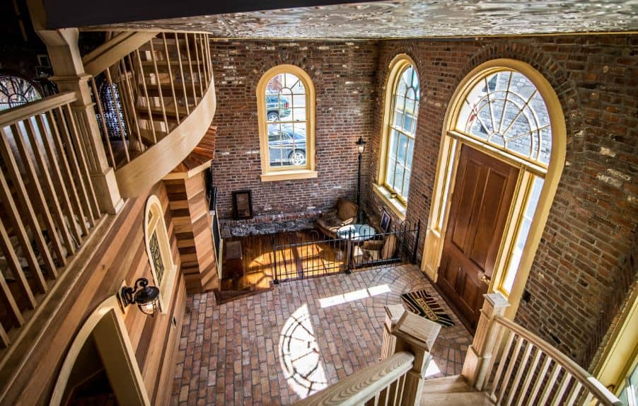 This charming industrial-style foyer has red bricks on its walls and floor that is illuminated by the side lights and arched transom window above the wooden main door that matches with the wooden stairs. Across from it is a wrought iron fence that separates the foyer from the living room.