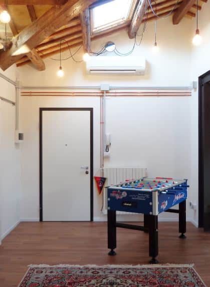 This simple foyer has a white door that blends with the white walls that contrast the hardwood flooring and wooden ceiling with exposed wooden beams. These are adorned with exposed wiring and tubing that line the walls and ceiling.