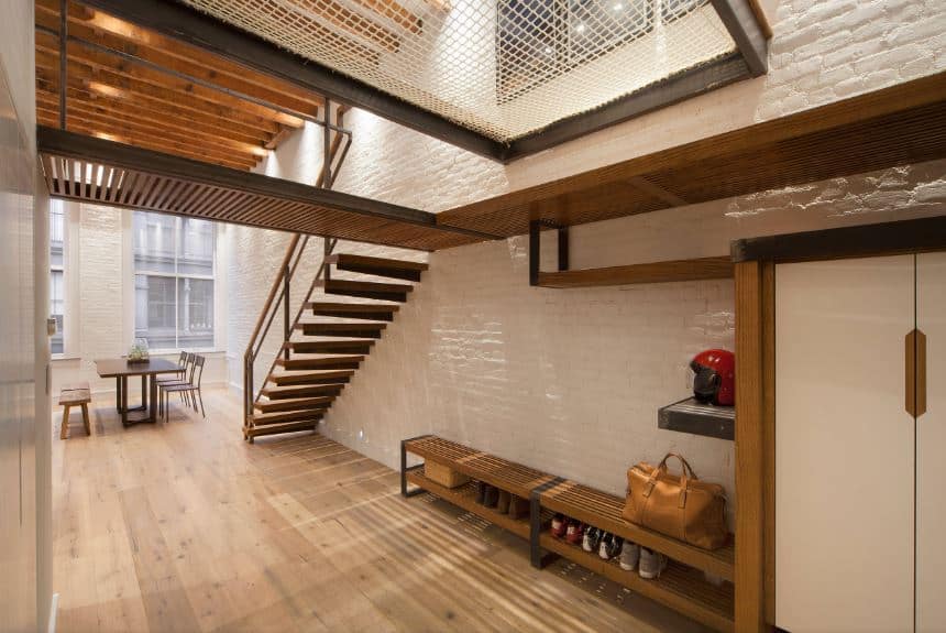 This industrial-style foyer has metal beams that support cat walks that would fit in a factory. This matches with the long wooden bench against the textured stone white wall with a lower shelf that serves as a shoe rack while the ones above are for helmets and hats.