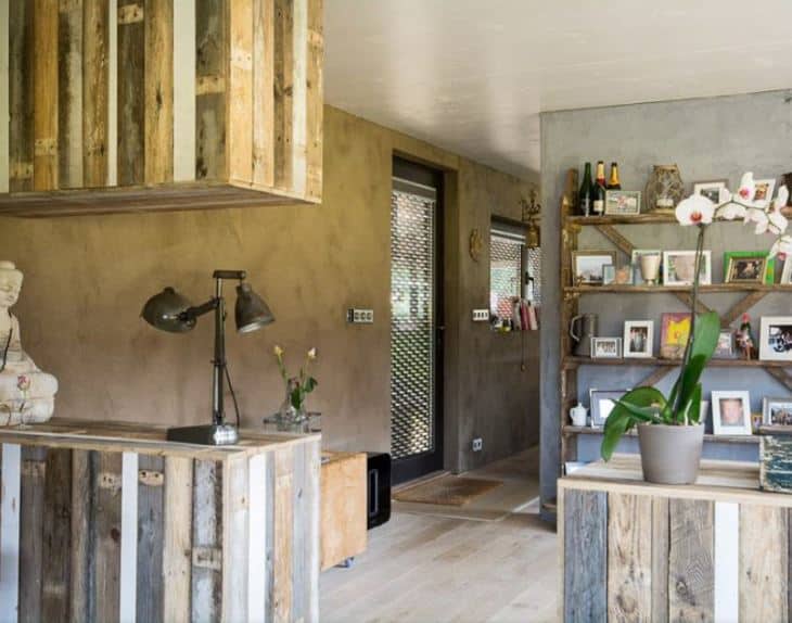 This foyer is a nice fusion of industrial-style elements and farmhouse-style elements. This foyer has a glass door with a peculiar industrial-style design to it. The door opens to a foyer with light hardwood flooring and industrial-style concrete walls that are complemented by various wooden structures.