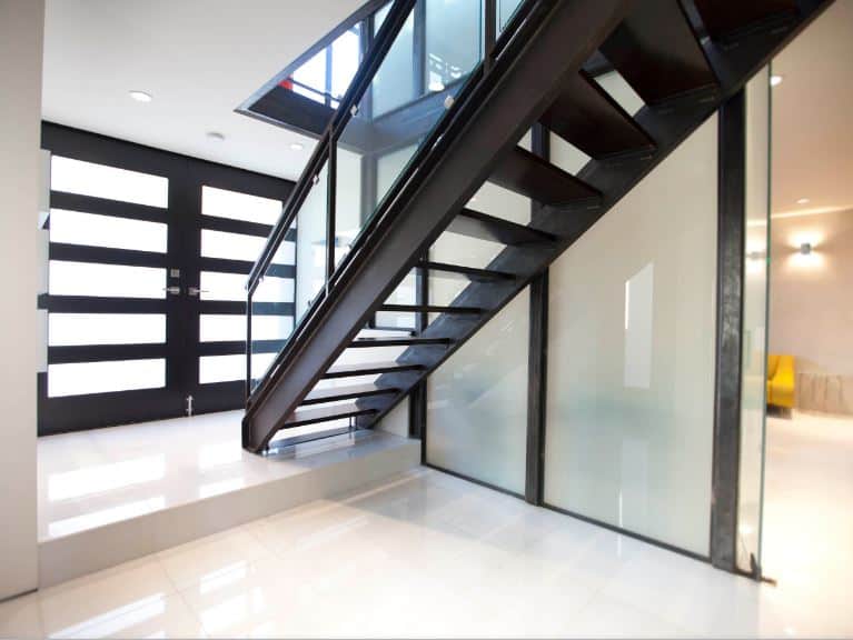 The highlight of this industrial-style foyer is the metal staircase that looks like it belonged in a factory. It has a dark metallic hue to it that matches with the dark hue of the main door with frosted glass panels that match the frosted glass walls of the staircase.