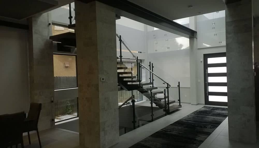 This foyer has exposed metal beams above it that matches the dark hue of the main door that has glass panels on it bringing in natural lights to the industrial-style flooring topped with a dark area rug running from the main door to the rest of the house with large stone pillars.