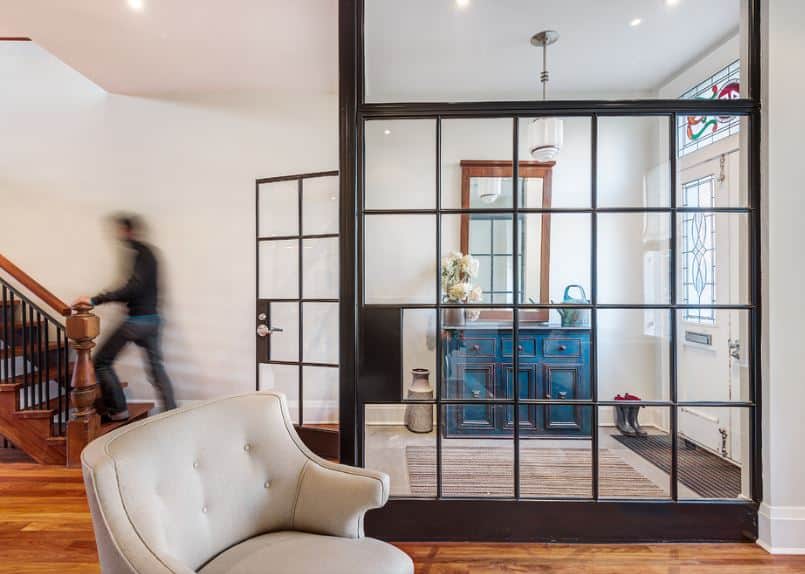 This charming industrial-style foyer is enclosed with glass walls and a glass door from the rest of the house. It has a white main door that has stained glass panels on it as well as in the transom window above that brings in natural lighting for the blue cabinet on the side.