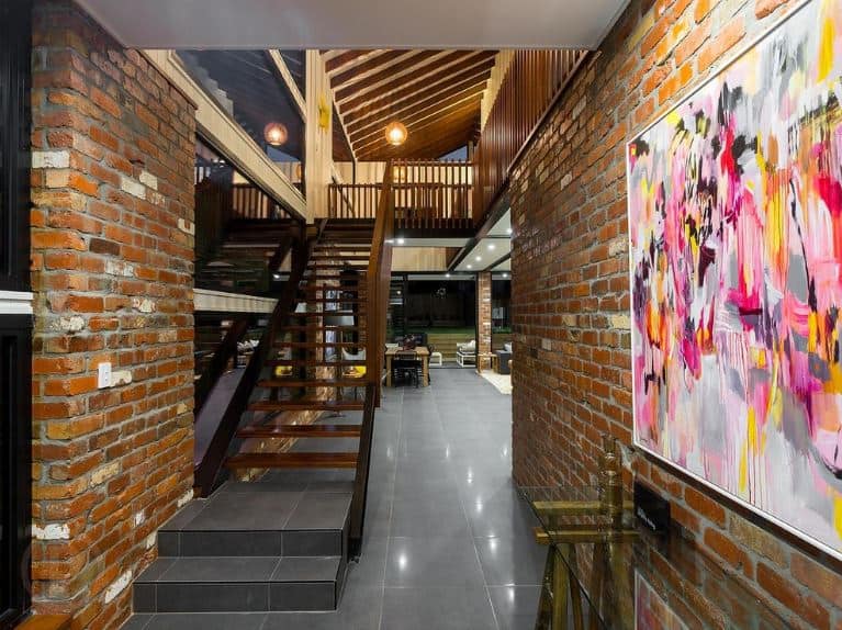 This small industrial-style foyer is like a warm hug filled with red brick walls that are adorned with a colorful abstract painting mounted above the glass-top console table with a brass frame that complements the dark gray tiles of the floor.
