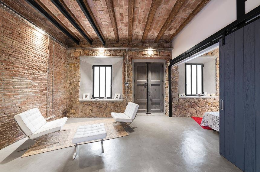 This industrial-style foyer has a lovely sitting area on the side that stands out against the red brick walls, stone walls and ceiling that has exposed wooden beams. All these earthy and textured surfaces are contrasted by the stark white modern sofas with a tufted finish.
