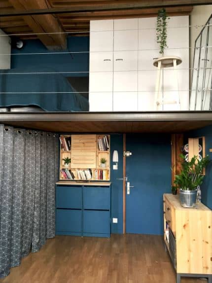 This foyer has a low wooden ceiling that also serves as the flooring of the loft above that has metal wire railings. This goes well with the blue main door that blends with the walls and the cabinet beside it that complements the dark hardwood flooring.
