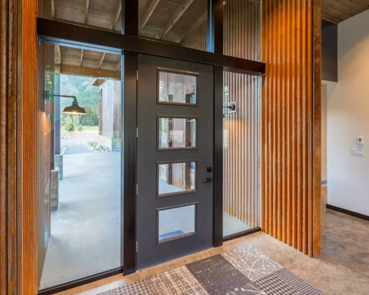This is an industrial-style foyer with rusty tin roofing material used on the walls adjacent to the gray main door that is framed with glass panels on three sides that bring in an abundance of natural lighting to the beige marble flooring topped with a gray patterned area rug.