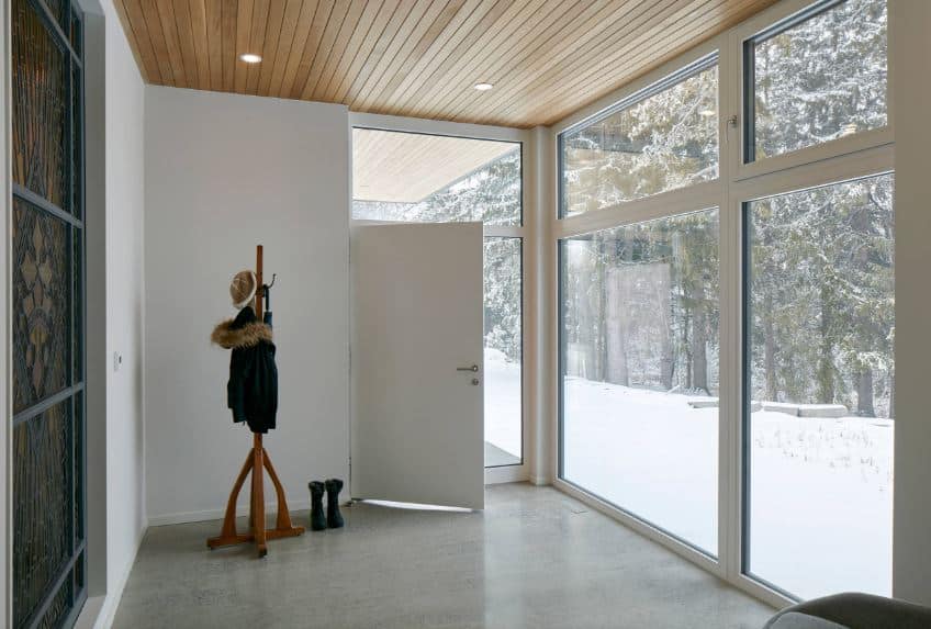 This industrial-style foyer has glass walls to showcase the beauty of the landscape scenery outside filled with snow and trees. The simple industrial-style flooring is adorned with a standing wooden rack for jackets and hats that pairs well with the wooden ceiling.