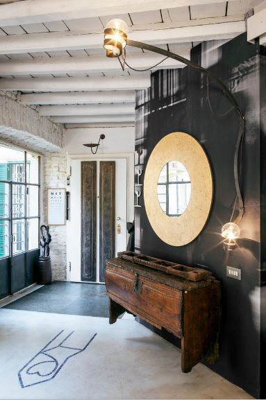 This foyer is almost filled to the brim with small details that solidify its industrial-style quality. It has a white wooden ceiling with exposed wooden beams that bleed into the black wall of the wall-mounted round mirror above a floating old wooden cabinet lit with a light bulb with exposed wires.