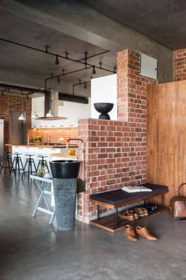 This simple and small industrial-style foyer has the same red brick walls and gray concrete flooring and ceiling as the rest of the house. On one side is a wooden bench with a black cushion and a shelf at the bottom of it to serve as a shoe rack.