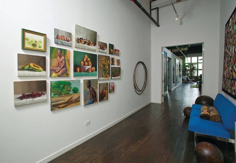 This hallway-like foyer has a dark hardwood flooring that matches the spherical decors beside the blue modern sofa that stands out against the white walls and ceiling with exposed pipes. Across from the sofa is a collection of wall-mounted paintings that bring color to the industrial-style foyer.