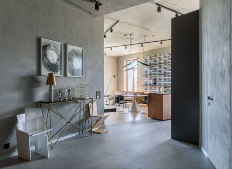 The gray main door that blends with the gray walls leads to this industrial style foyer that has concrete flooring and a modern console table bearing decors and a table lamp. The two paintings mounted on the concrete wall above the table adds to the aesthetic.