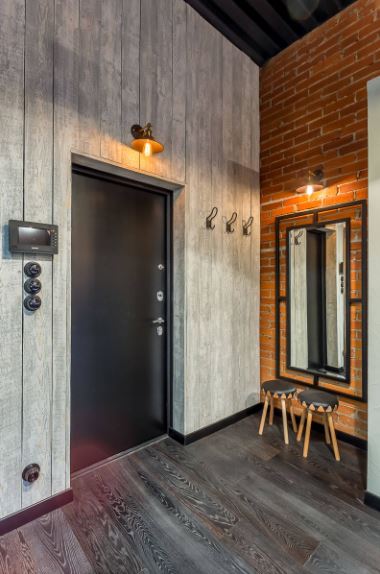 The black main door is lit with a warm yellow light by the industrial-style wall lamp above the door. This matches with the wall lamp above the mirror that is mounted on the adjacent wall that is made of red bricks and paired with a couple of small wooden stools.