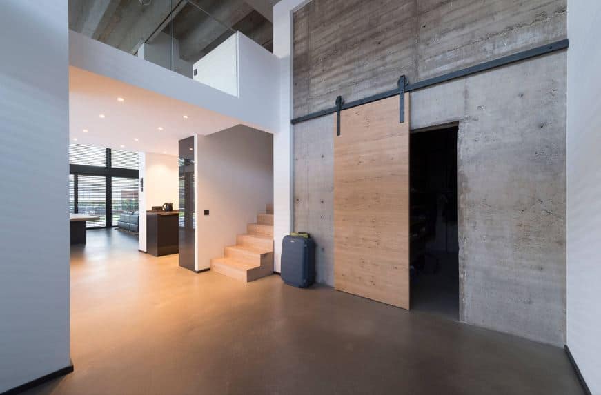 This wide industrial-style foyer has concrete flooring that pairs well with the concrete wall that supports the iron rung of the wooden sliding door that matches with the wooden stairs leading to the second level that has glass railings.