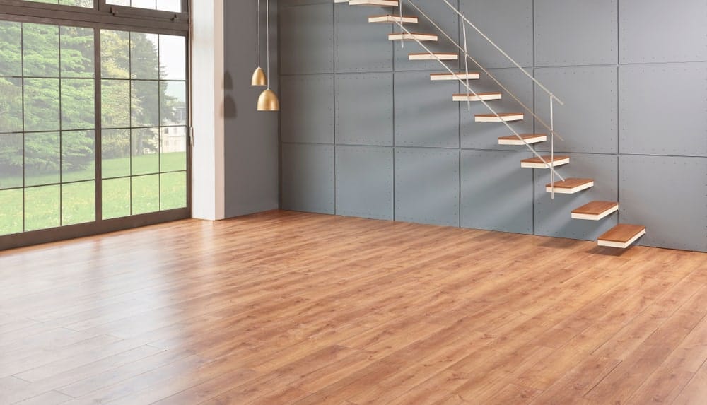 This wide foyer has a tall glass wall with gray frames that match the gray industrial walls that support the floating stairs with the same wooden hue to the steps as the hardwood flooring that is illuminated by the natural lights coming in from the glass walls.