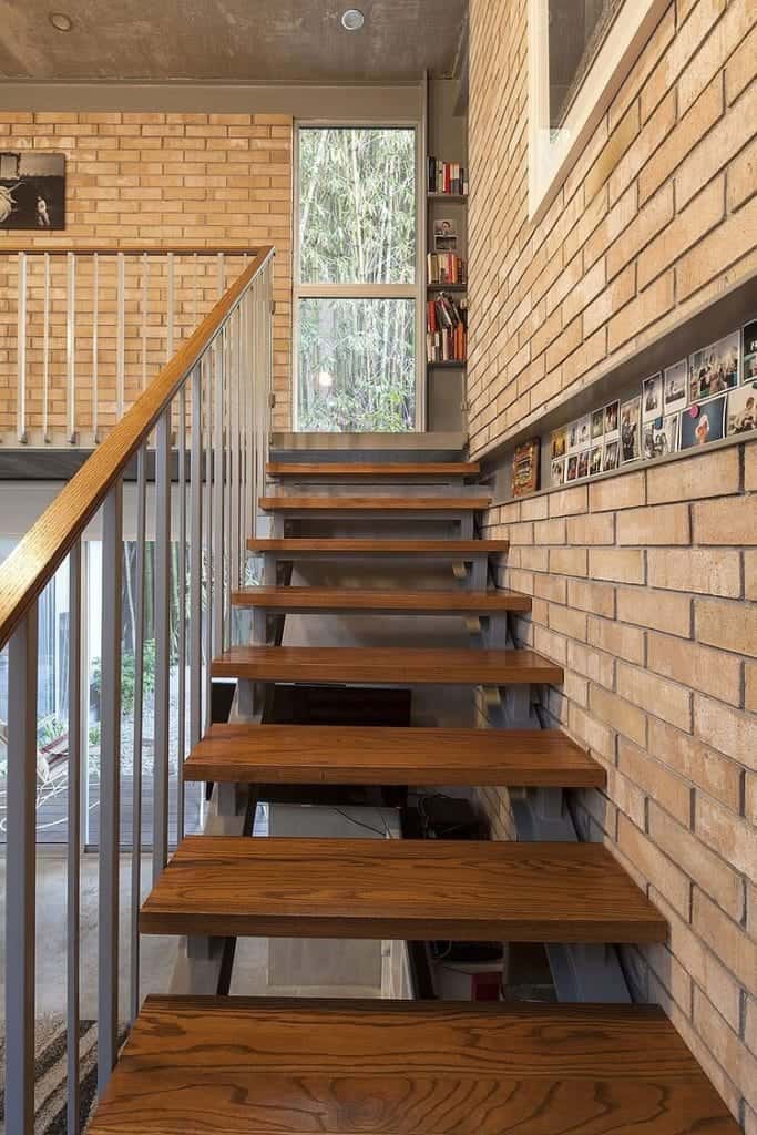 The glass door leads to this industrial-style foyer that has red brick walls adorned with photos and a gray concrete ceiling that has recessed lights. Right beside the door is a small bookshelf filled with books and decors that adds a bit of homey quality to the foyer.