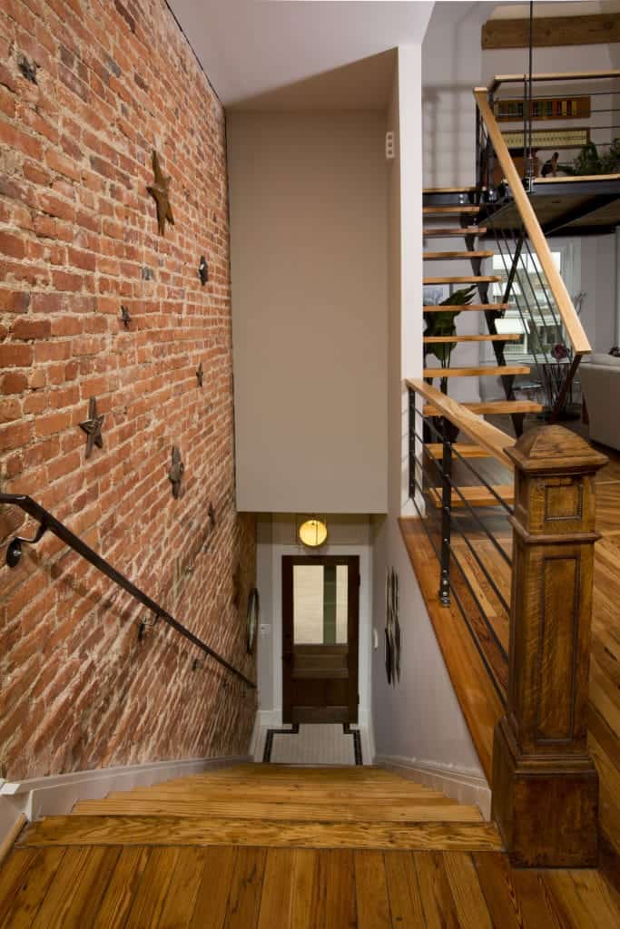 This industrial-style foyer has a wooden main door with a glass panel on it lit by an industrial-style wall lamp above. The small foyer directly leads to a wooden stairs that has a red brick wall on one side and white wall on the other side leading up to the rest of the house.