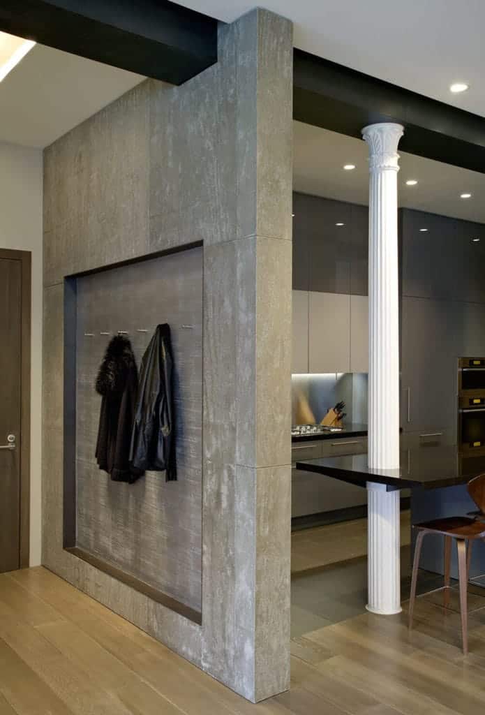 This foyer is separated from the kitchen by a gray concrete wall that has an alcove with hooks that can be used as coat racks and hat racks for those who enter the brown wooden main door that contrasts the beige walls but complements the hardwood flooring.