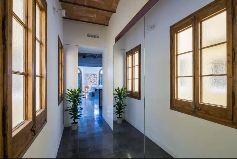 This narrow hallway-like foyer has a large seamless mirror on the right side that gives it an illusion of being bigger than reality. It has industrial-style gray flooring tiles that contrast the white walls that has a pair of wood-framed windows with frosted glass.