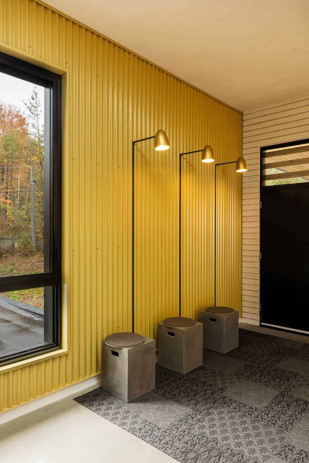 This simple Industrial-style entry hall has a gray patterned area rug on its off white flooring. This is brightened by the delightful yellow hue of the walls made of metal and the wall-mounted lamps attached to it over three small built-in concrete stools.