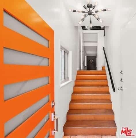 Upon entry of this bright orange front door that has frosted glass panels, you are welcomed with an industrial-style foyer that is narrow and has white walls and ceiling with an industrial-style chandelier above the terracotta stairs leading up to the rest of the house.