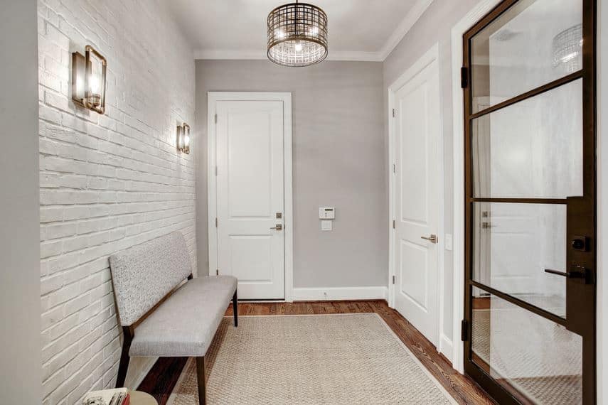 This simple industrial-style foyer has a textured white wall with a brick wall pattern adorned with a couple of wall-mounted lamps that match with the round pendant light from the light gray ceiling matching the walls and woven area rug as well as the cushioned bench.