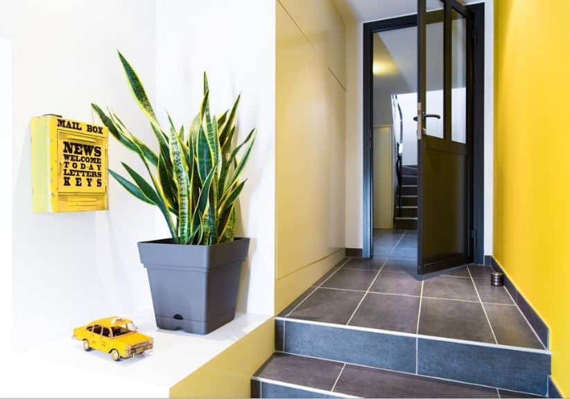 This foyer has a cheerful welcome for the guests with its yellow wall that matches with the mailbox and the taxi decor beside the potted plant. This yellow wall is contrasted by the matte industrial-style flooring as well as the black wooden door.