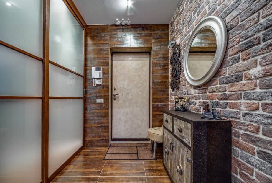 This foyer is dominated by the earthy hues of the brick walls and the brown tiles of the flooring that extends to the wall framing the main door. Adjacent to this is the brick wall that has a wooden cabinet and mirror. Across from this is a frosted glass wall.