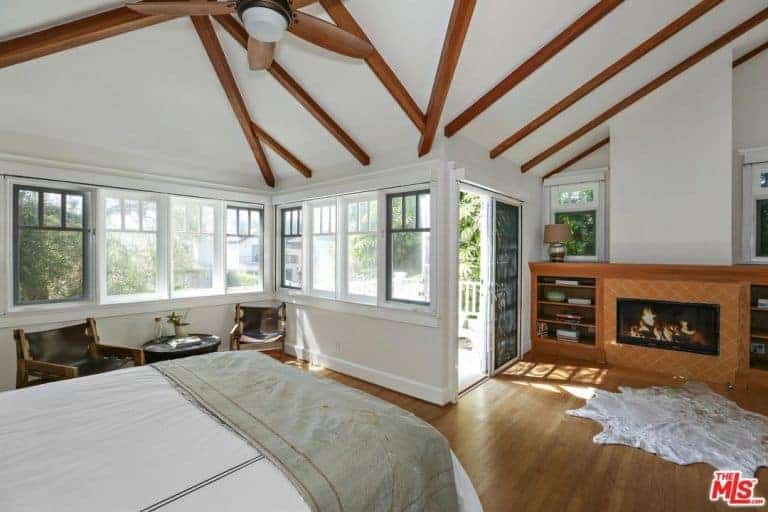 Spacious primary bedroom featuring hardwood flooring and a white ceiling with exposed beams. The room has a set of seats on the side with a small coffee table and a fireplace with built-in shelves on both sides.