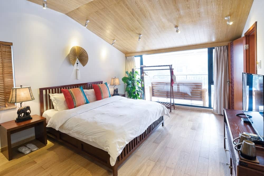 Cozy primary bedroom with a wooden bed and nightstands topped with elephant table lamps. It is illuminated by white track lights that are mounted on the vaulted ceiling clad in light wood planks.
